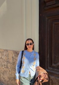 Femme portant des lunettes de soleil noires et un cardigan bleu à motif losanges se tient contre un mur beige, tenant une veste en cuir marron et un sac noir.