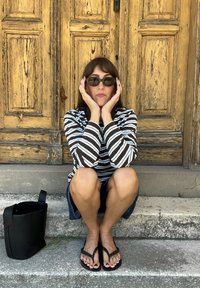 Camisa de manga longa às riscas em preto e branco, calções de ganga, chinelos pretos. Uma mala de tote preta está ao lado de uma pessoa sentada nos degraus.