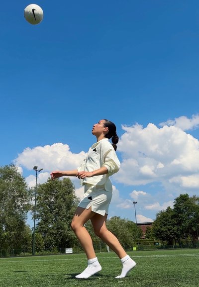 Un balón de fútbol blanco de Nike en el aire sobre una persona que lleva un atuendo deportivo ligero beige con acentos negros, de pie sobre la hierba verde bajo un cielo azul.