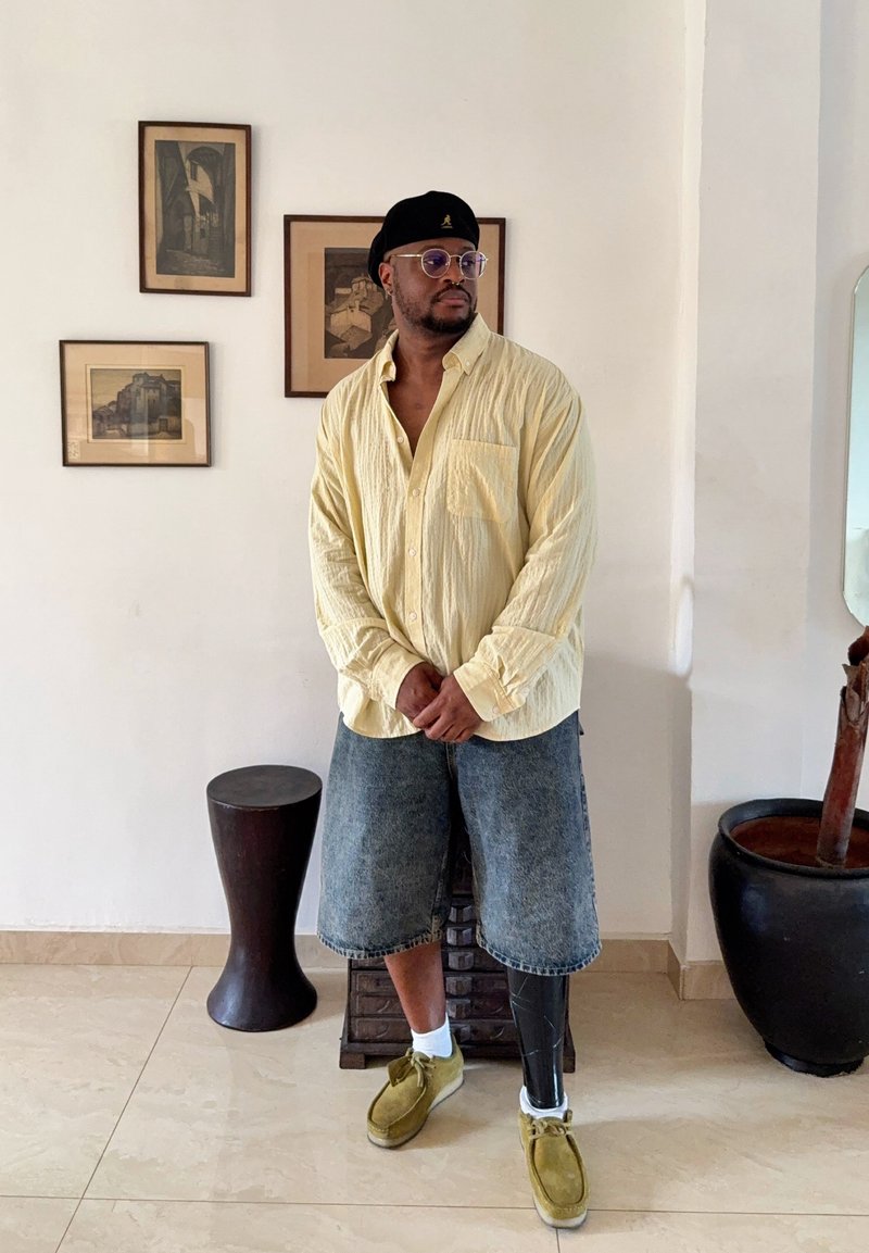 Homme portant une chemise boutonnée jaune clair, un short en jean, une prothèse noire à la jambe, des chaussures vertes et un béret noir, debout dans une pièce minimaliste avec des œuvres encadrées.