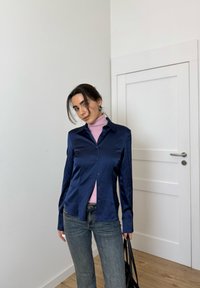 Temno modra satenasta srajca z zaponkami in razširjenim ovratnikom, kombinirana s svetlo roza puloverjem z visokim ovratnikom. Nošeno z modrimi jeansa in črno torbico.