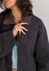 Donna con capelli scuri di lunghezza fino alle spalle, che indossa una giacca scura con colletto in velluto a coste marrone, tenendo la giacca vicino alla zip con la mano sinistra.