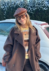 Jeune femme aux longs cheveux blonds portant un manteau marron, un pull à rayures, une casquette en cuir et de grandes lunettes de soleil, souriant debout à l'extérieur près d'une voiture recouverte de neige.