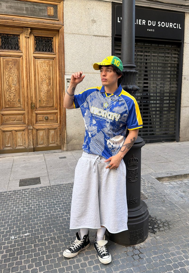 Jeune homme portant un t-shirt bleu et jaune, un short long gris, des baskets montantes noires et une casquette verte et jaune, debout près d’un lampadaire dans une rue en ville.