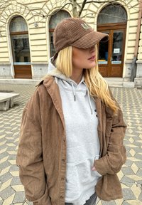 Chaqueta de camisa de pana marrón sobre un sudadera gris, con cordón ajustable, complementada con una gorra marrón a juego que muestra un bordado verde.