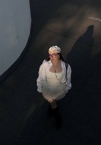 Camisa branca oversized sobre um vestido branco justo com saias de babados, combinada com botas pretas e um lenço de cabeça estampado. Os acessórios incluem um colar de pérolas.