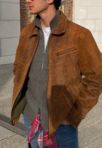 Homme portant une veste en cuir marron sur un sweat à zip gris et un jean bleu, avec une chemise à carreaux rouge nouée à la taille, debout à l'extérieur près d'un mur en briques.