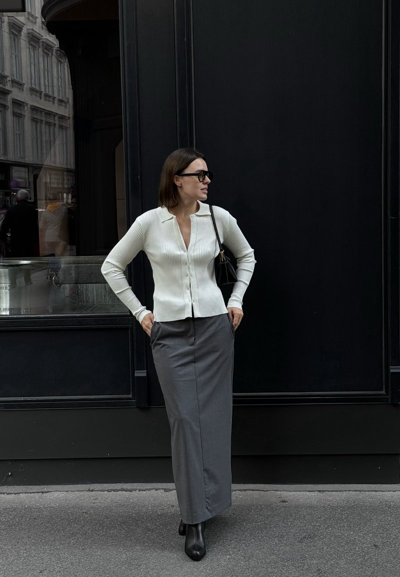Λευκό χ ribbed cardigan με γιακά, γκρίζα μακριά φούστα και μαύρες μπότες, συνοδευόμενο από μαύρη τσάντα, ποζαρισμένο μπροστά σε σκοτεινό φόντο.