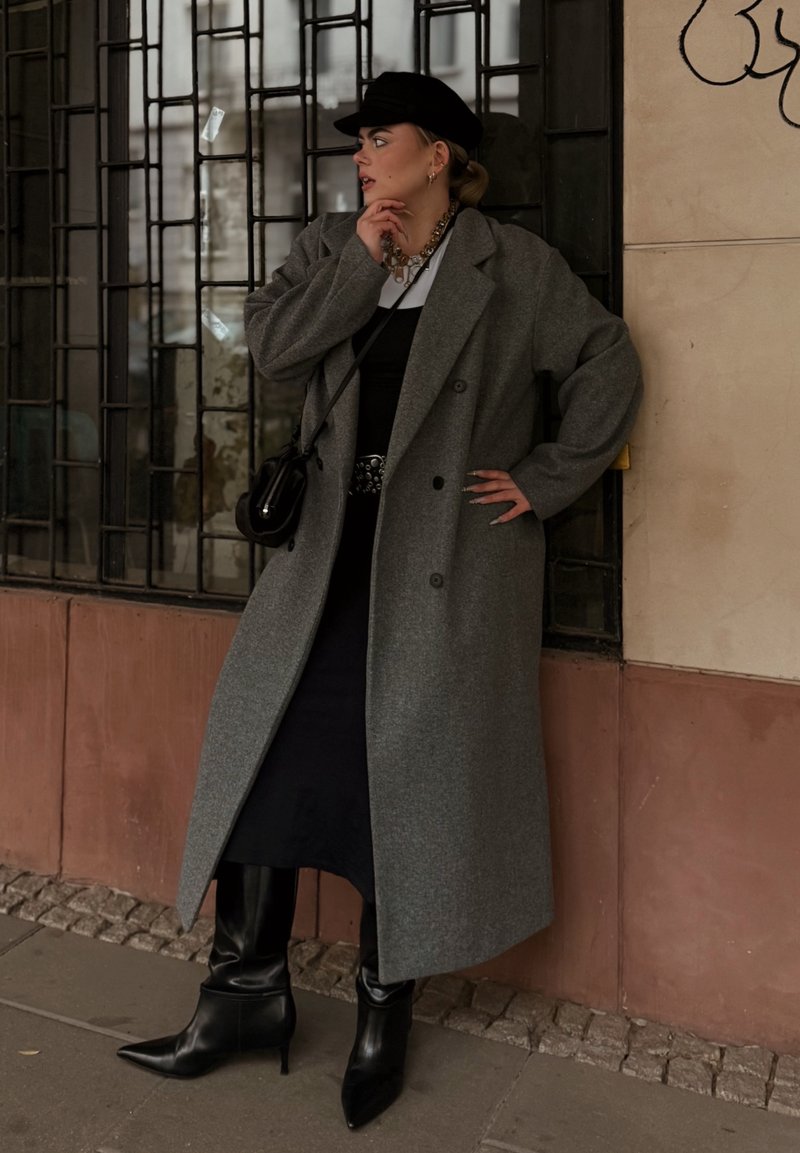 Grauer Wollmantel mit weitem Revers, schwarzes tailliertes Kleid, auffälliger Gürtel, schwarze kniehohe spitze Stiefel und eine schwarze Mütze auf einer Straße in der Stadt.