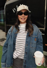 Femme portant des lunettes de soleil, une casquette beige, une veste en denim et une chemise rayée, tenant une tasse de café en plein air dans un cadre urbain.