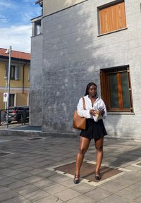Chemise blanche à boutons, jupe plissée noire et sac en cuir marron. Debout sur un pavé en pierre, en train de lire un magazine. Décor urbain.