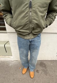 Personne portant une veste bomber vert olive, un jean bleu et des chaussures en daim beige debout sur un sol en béton près d'un mur de couleur crème.