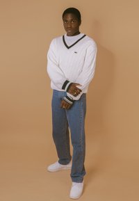 Weißer Kabelstrickpullover mit V-Ausschnitt und dunkelgrünem Besatz, kombiniert mit blauen Jeans und weißen Sneakers. Vor einem beigen Hintergrund.