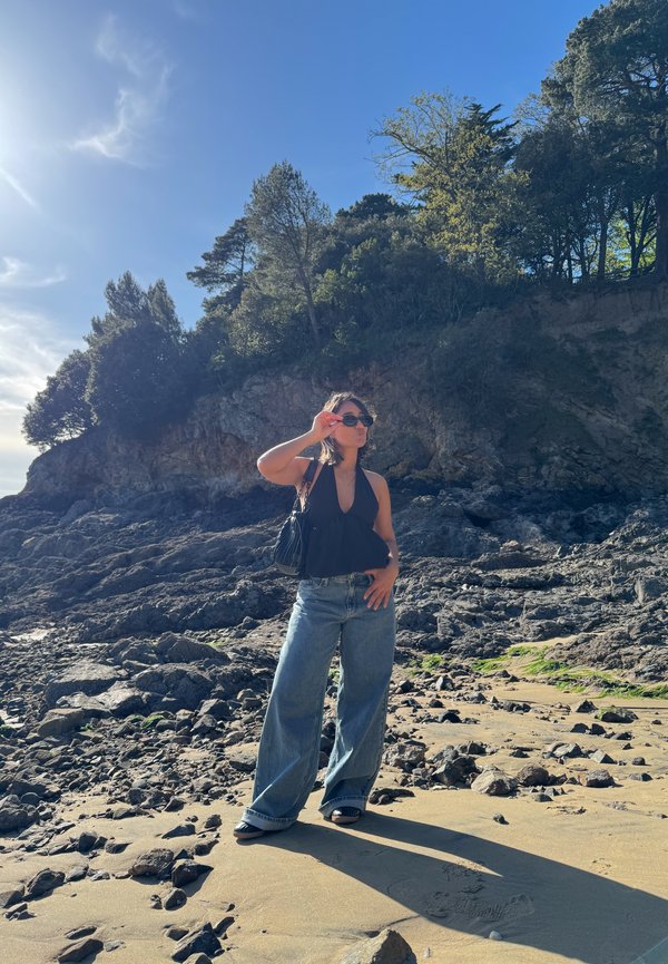 Vrouw met zonnebril en zwart topje staat op een rotsachtig strand met bomen op de kliffen onder een helderblauwe lucht, de zon schijnt van links.
