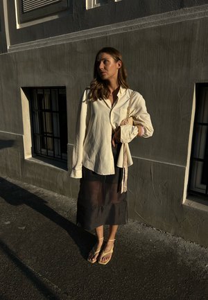 Mujer de pie contra una pared gris con dos ventanas con barrotes, vestida con una camiseta color crema, una falda negra transparente, sandalias amarillas y llevando un bolso beige.