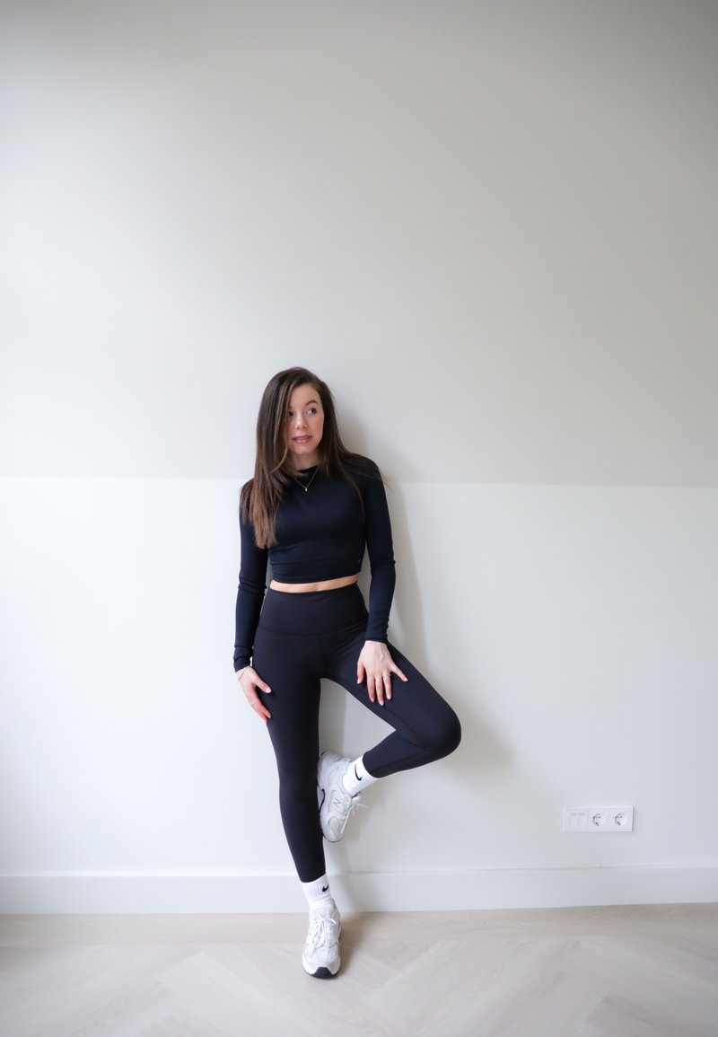 Μαύρο, μακρυμάνικο crop top και ψηλόμεσες κολάν σε συνδυασμό με λευκά αθλητικά παπούτσια; επίπεδη τοίχος με φωτεινό χρώμα δαπέδου.