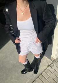 Μαύρο σακάκι, λευκό ριμπ tank top, λευκά σορτς, μαύρες μπότες μέχρι το γόνατο με αγκράφες, λευκές κάλτσες. Στέκεται σε μια τσιμεντένια επιφάνεια.