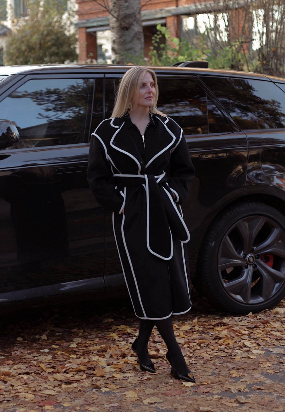 Black coat with white piping, belted at the waist, long sleeves, and knee-length. Black high heels, standing beside a black SUV on a leaf-covered ground.