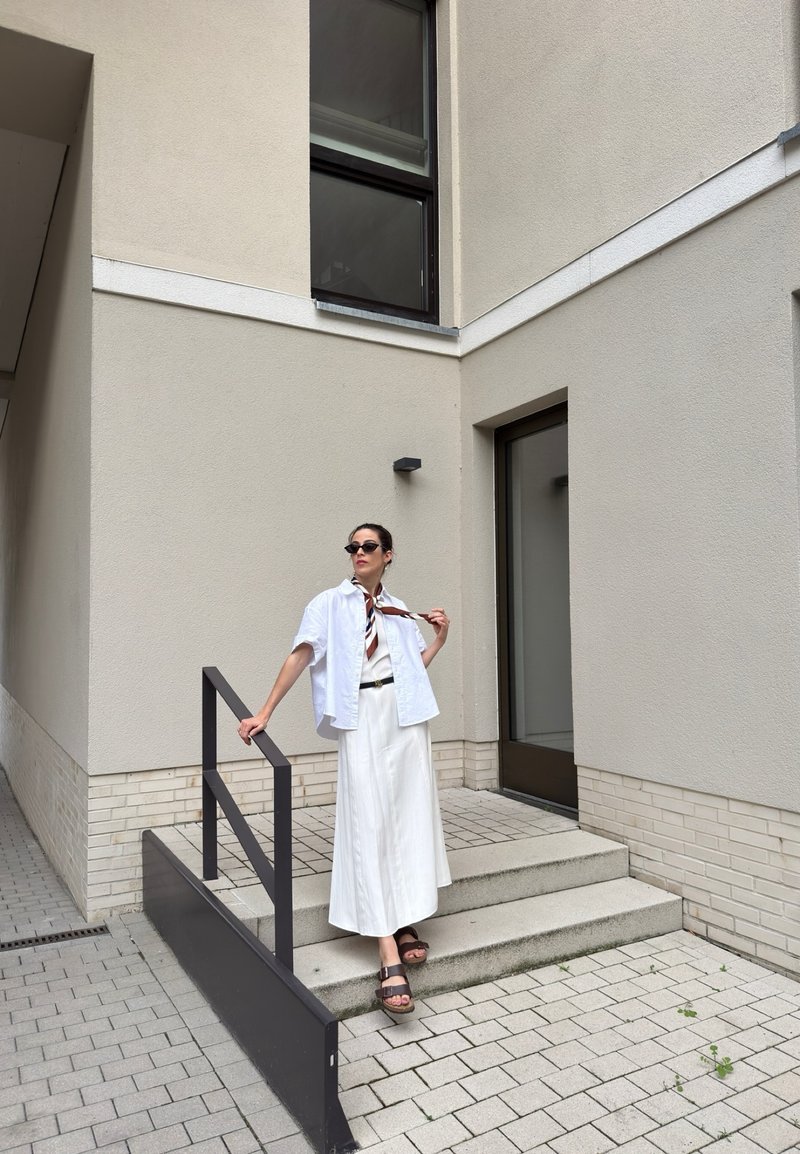 Woman wearing white skirt and shirt stands on outdoor stairs next to beige building wall holding black railing and adjusting scarf.