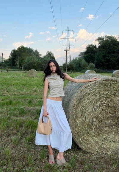 Mujer con top beige y falda blanca sosteniendo un bolso marrón claro, de pie con una mano sobre un gran fardo de heno en un campo con hierba, árboles y líneas eléctricas.