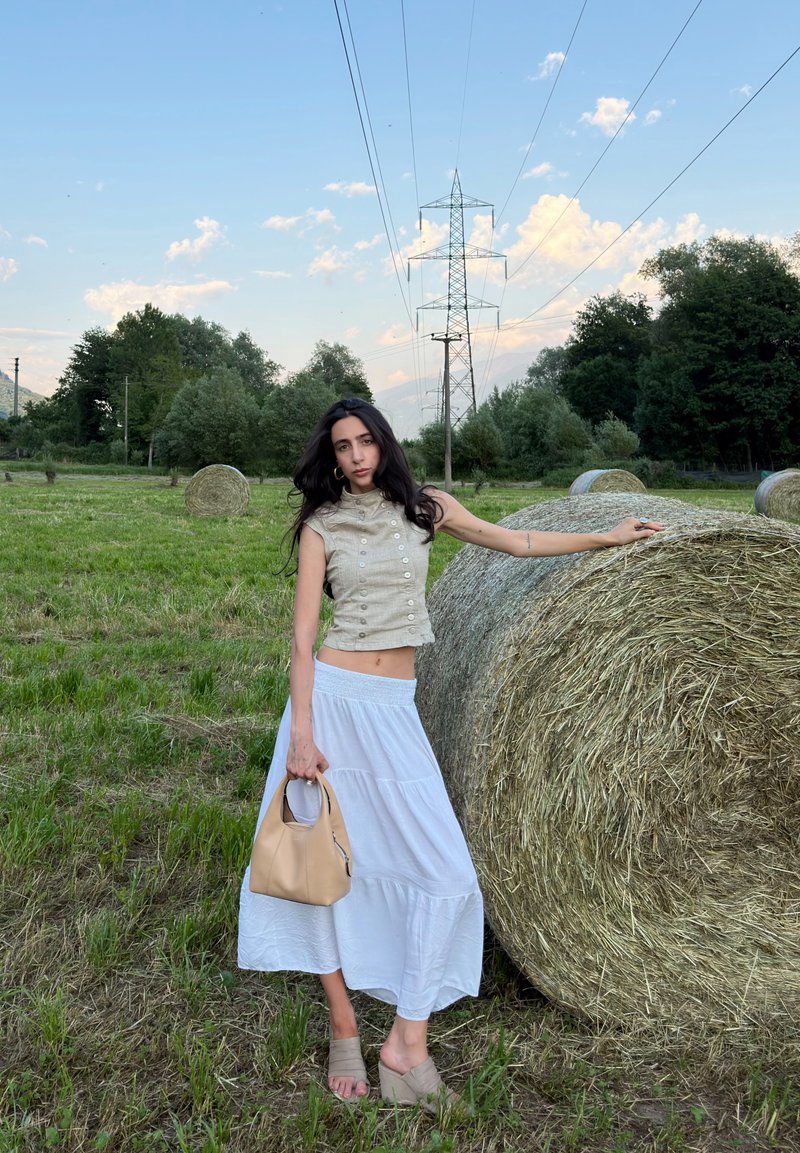 Femme en haut beige et jupe blanche tenant un sac beige, debout avec une main sur une grosse botte de foin dans un champ herbeux avec des arbres et des lignes électriques.