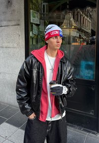 Jeune homme en veste en cuir, sweat à capuche rouge et bonnet tenant une tasse de café, debout sur un trottoir en ville près d'une vitrine en verre au crépuscule.