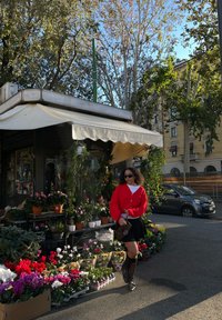Gilet rouge par-dessus une chemise blanche, jupe noire ; magasin de fleurs avec des fleurs colorées en pots, feuillage vert, et une auvent.