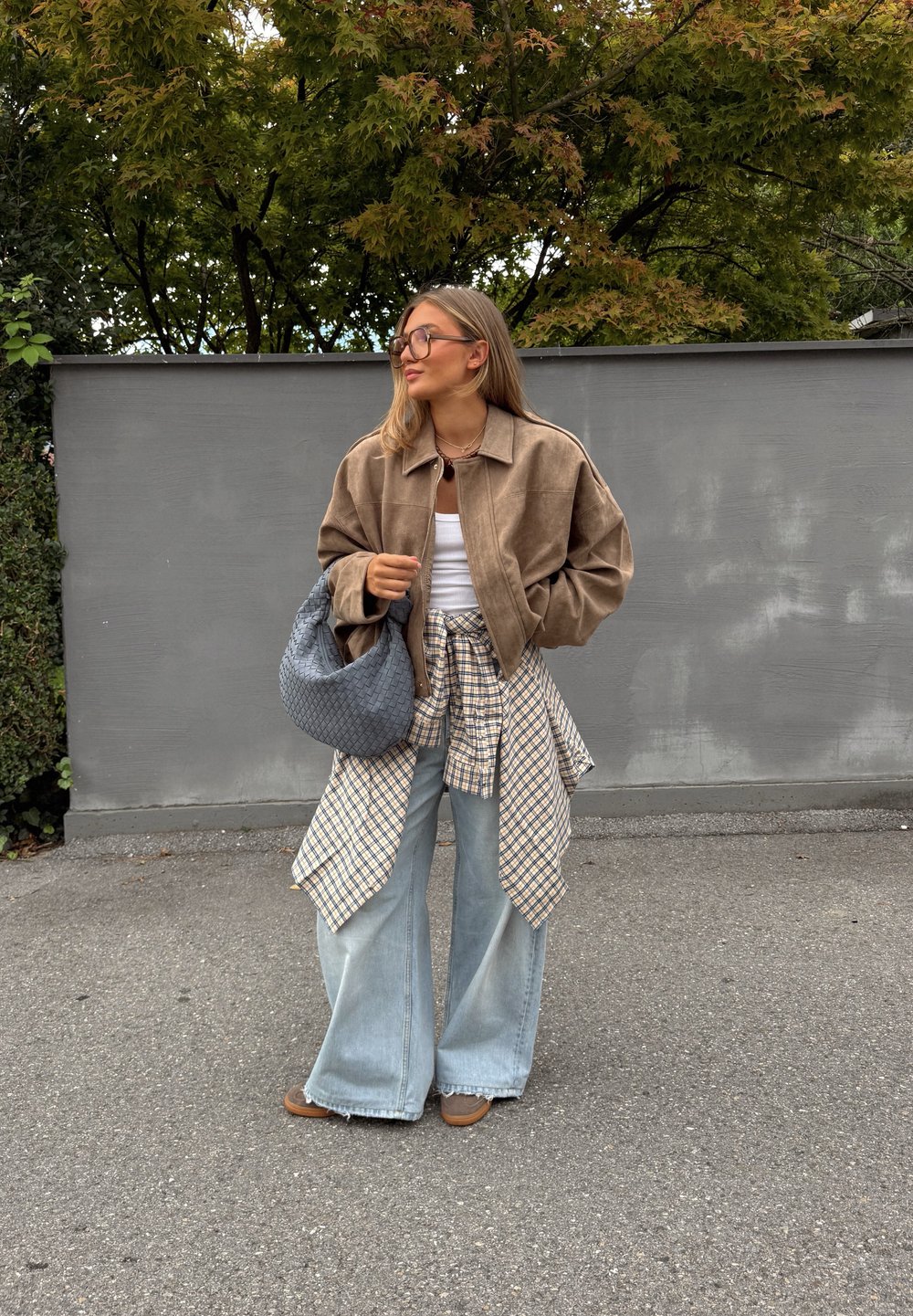 Brun semsket cropped jakke, hvit tanktopp, rutete skjorte knyttet i midjen, vide blå jeans, lys brune sko og en grå vevet håndveske.