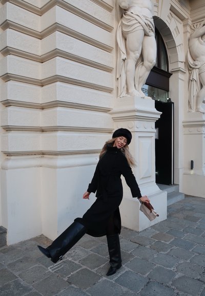 Abrigo negro con cinturón, botas de cuero negras hasta la rodilla y un sombrero negro circular. Ella sostiene una pequeña bolsa de papel mientras posa frente a un edificio.