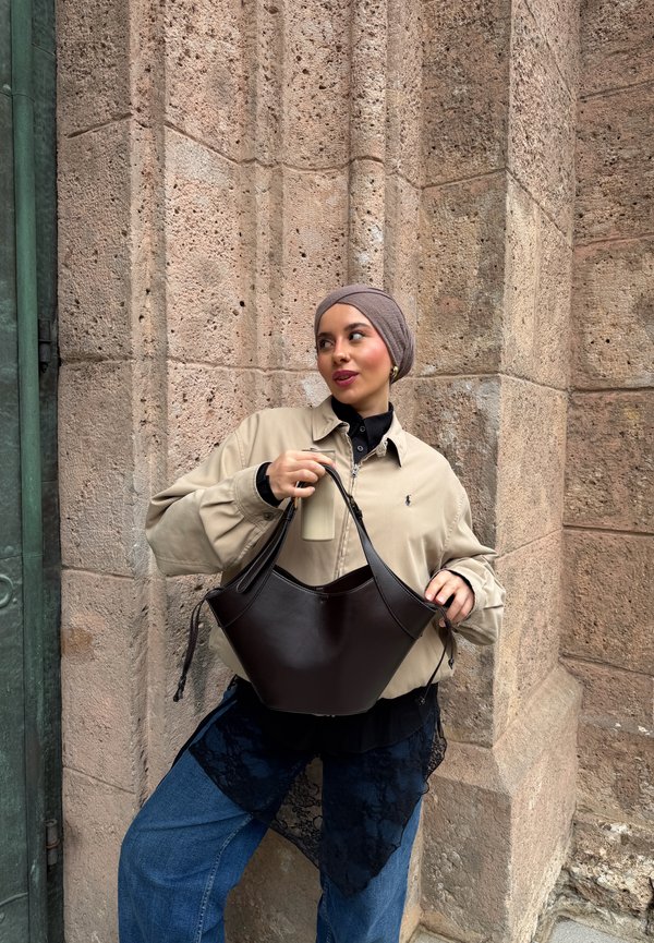 Mujer con chaqueta beige y pañuelo en la cabeza sostiene un bolso de cuero negro y una botella beige, de pie contra una pared de piedra con textura al aire libre.