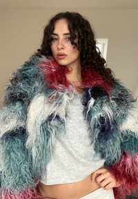 Manteau en fourrure coloré aux nuances variées de bleu, rose et blanc. Matière texturée et hirsute avec une coupe ample. Porté par-dessus un haut gris.