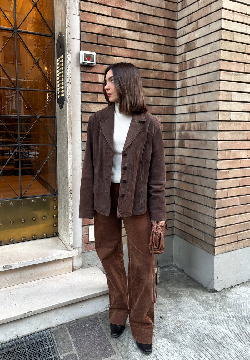 Brown corduroy blazer with buttons, paired with matching wide-leg trousers. White roll-neck underneath, holding brown gloves. Textured brick background.