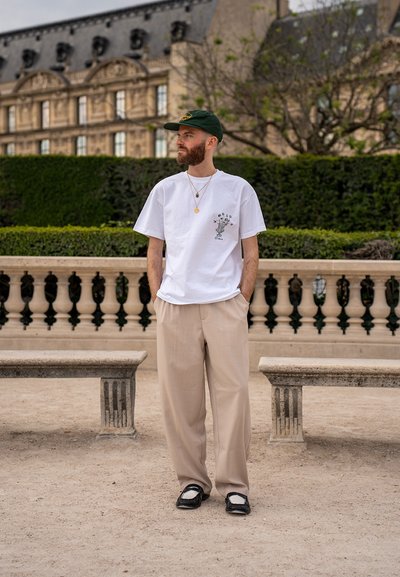 Camiseta blanca de algodón con diseño impreso, pantalones de tela beige con plisados, sandalias en blanco y negro, gorra verde y collares de oro en capas.