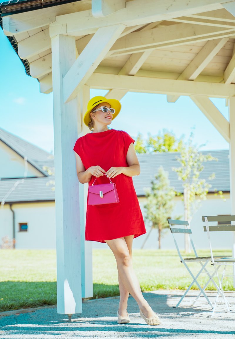 Femme en robe rouge et chapeau jaune se tenant sous une pergola en bois blanc, tenant un sac à main rose, avec des chaises d'extérieur et de la verdure en arrière-plan.