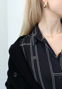 Camisa negra de botones con rayas verticales blancas, puesta debajo de un abrigo negro. Pendientes dorados en forma de lágrima y un delicado collar dorado con un colgante.