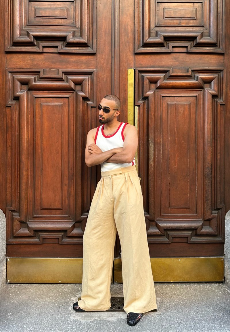 Homme portant des lunettes de soleil, un débardeur blanc avec finition rouge et un pantalon large beige, debout les bras croisés devant de grandes portes en bois ornées.