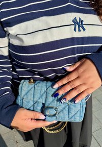 Manos con uñas de punta francesa en azul marino y nude, una con el logo blanco de NY, sosteniendo un bolso de denim acolchado; la persona lleva un suéter a rayas en azul marino de NY.