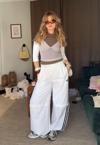 Bílé, volné tepláky s černými bočními pruhy, spárované s topem ve stylu color-block (hnědá a světle fialová) a slunečními brýlemi.