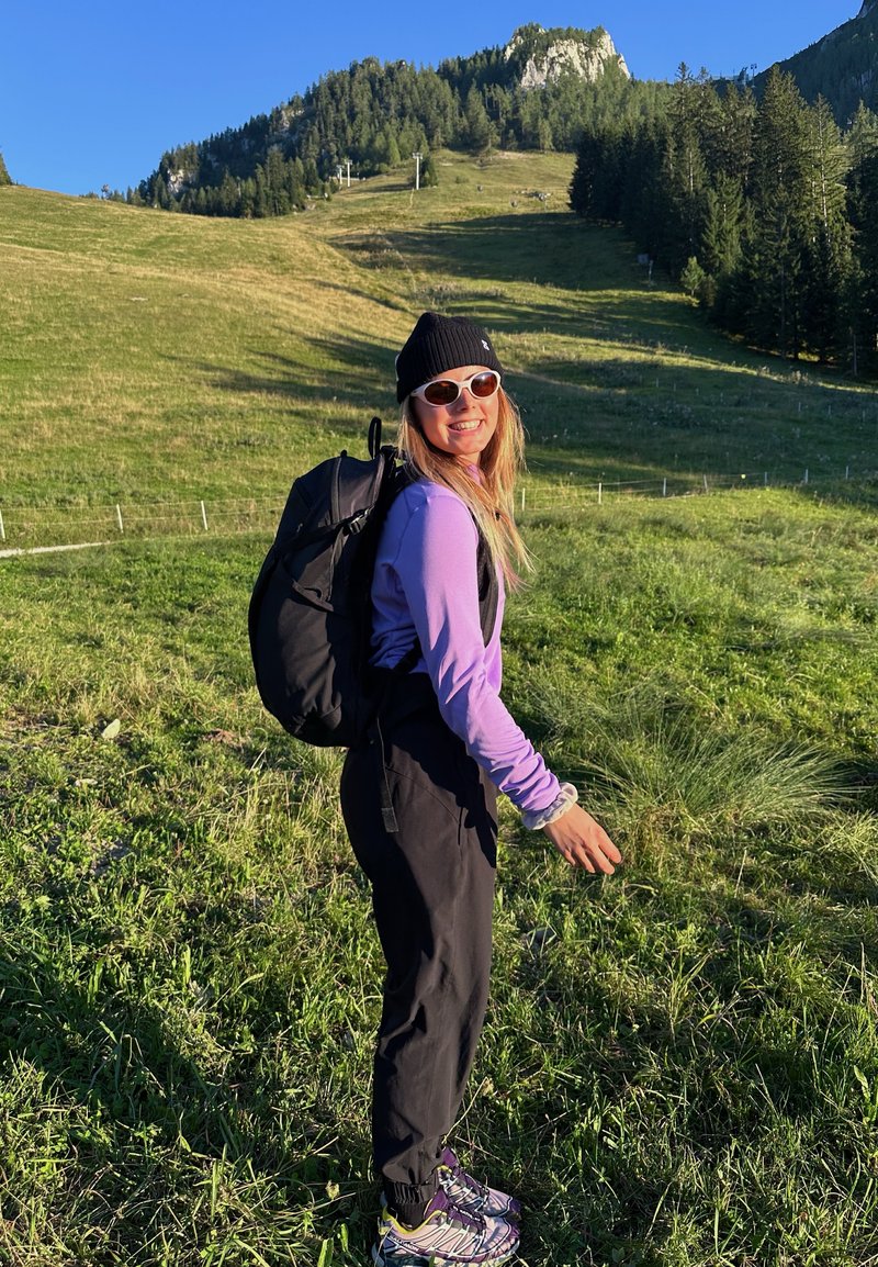 Femme en chemise à manches longues violette et pantalon noir, portant des lunettes de soleil et un bonnet noir, avec un sac à dos noir. Fond de gazon vert.