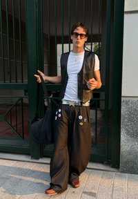Gilet en cuir noir porté sur un t-shirt blanc, pantalon noir large avec divers boutons, portant un sac noir, chaussures marron, debout près de portes vertes.