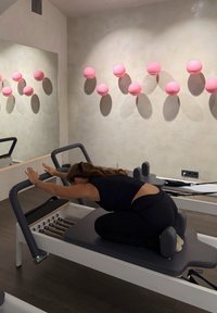 Reformer Pilates-maskine med en grå polstret platform og sorte modstandsbånd. Lyserøde runde vægdekorationer og et stort spejl i et lyst rum.