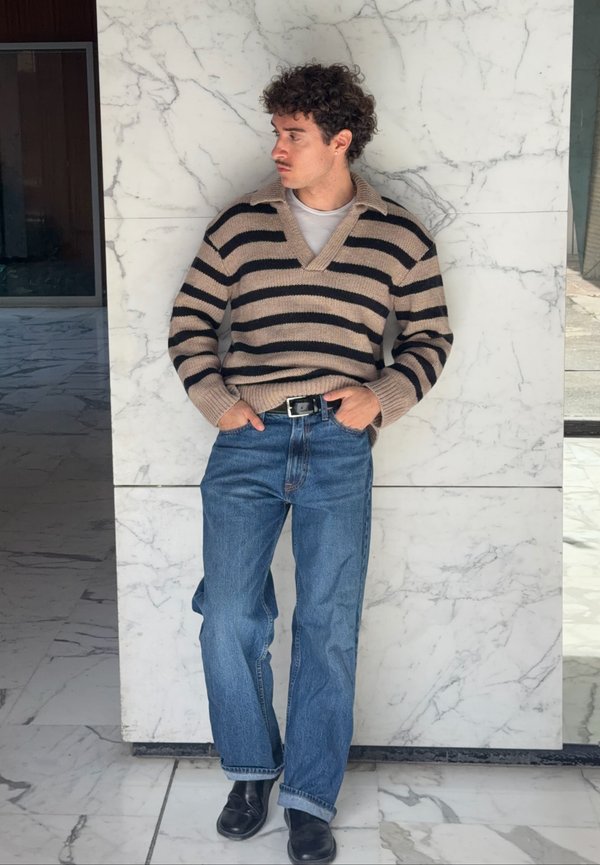 Hombre con cabello rizado que lleva un suéter de rayas marrón y negro, vaqueros azules y zapatos negros, de pie contra una pared de mármol.