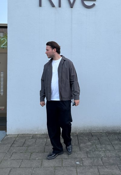 Chaqueta ligera gris sobre una camiseta blanca, combinada con pantalones anchos negros y zapatos negros, de pie sobre una superficie pavimentada contra una pared.