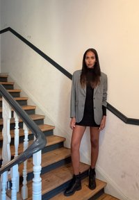 Blazer cinza sobre um vestido preto, de pé em escadas de madeira. Botas de combate pretas, textura suave. Corrimão branco e parede neutra ao fundo.