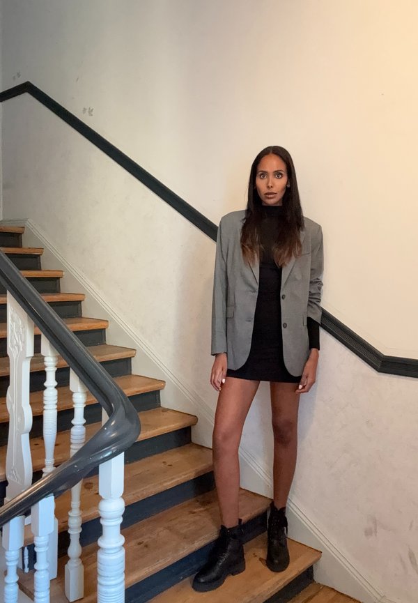 Blazer gris sobre un vestido negro, de pie en unas escaleras de madera. Botas de combate negras, textura suave. Barandilla blanca y pared neutral de fondo.