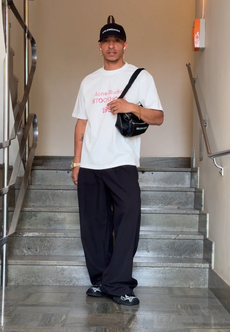 Homme debout dans un escalier intérieur portant un t-shirt blanc graphique, un pantalon large noir, une casquette Balenciaga noire et portant un sac à bandoulière Balenciaga noir.