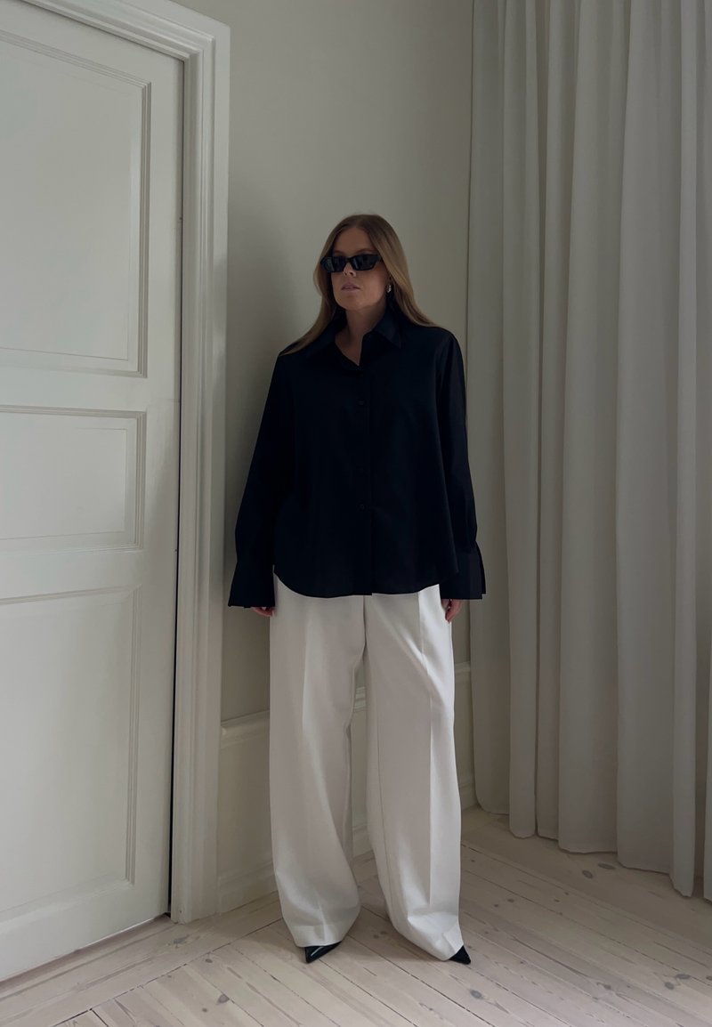 Femme portant une chemise noire oversize, un pantalon blanc à jambes larges, des chaussures pointues noires et des lunettes de soleil foncées, debout à l'intérieur près d'une porte et d'un rideau.