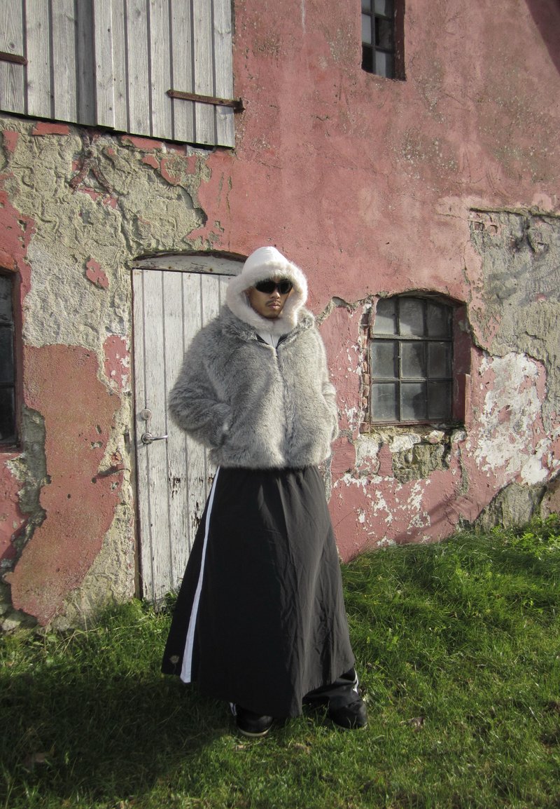 Giacca in pelliccia ecologica grigia con cappuccio bianco, abbinata a una lunga gonna nera. Lo sfondo presenta un muro rosa consumato e una porta di legno.