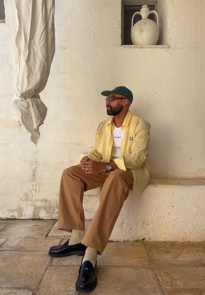 Hombre con barba que lleva una camisa amarilla, camiseta blanca, pantalones marrones, gorra verde y zapatos negros sentado en un borde de piedra contra una pared beige con un jarrón.