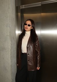 Γυναίκα με μακριά σκουρόχρωμα μαλλιά που φοράει μαύρα γυαλιά ηλίου, λευκό ζιβάγκο, σκούρο καφέ δερμάτινο μπουφάν και μαύρο παντελόνι, στέκεται δίπλα σε έναν ανελκυστήρα.